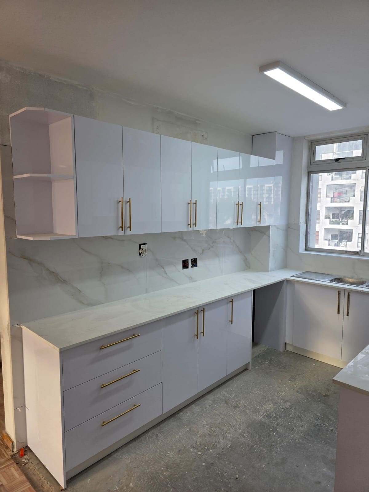 The same kitchen after: white high-gloss wall and base cabinets with brushed gold handles, a white marble-look splashback and worktop, and the sink run under the window.