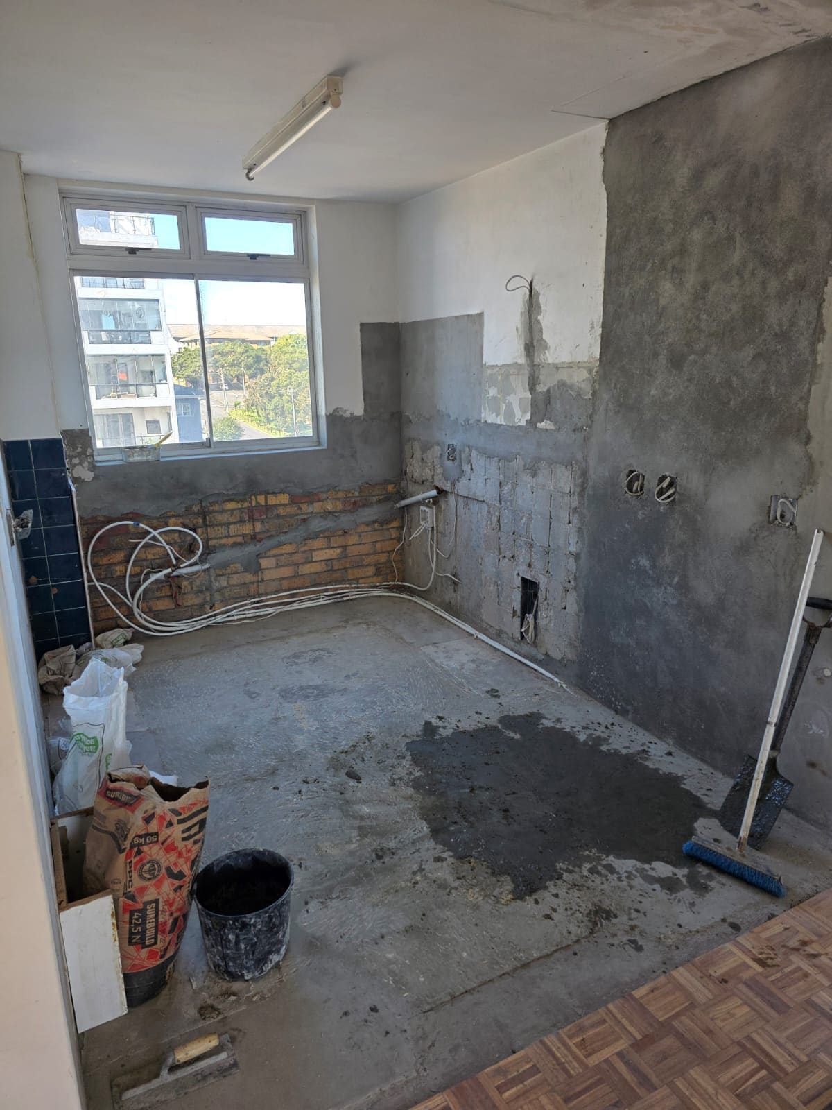 The kitchen before: a stripped room with bare brick and plaster walls, exposed plumbing under the window, a concrete floor and building materials.