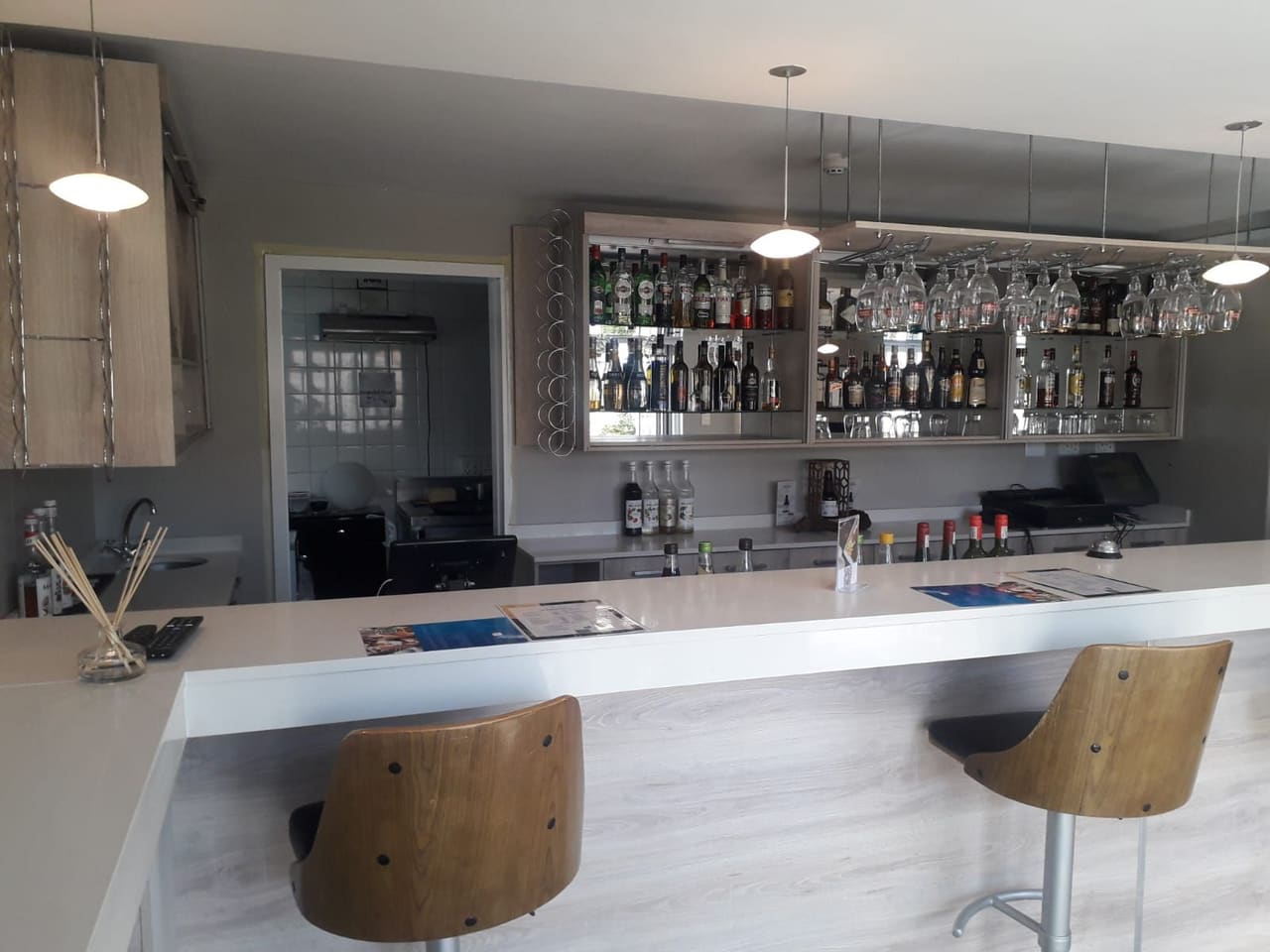 Bespoke bar counter with a white top and timber-look front, in front of a back bar of lit open shelving and a suspended glass rack.
