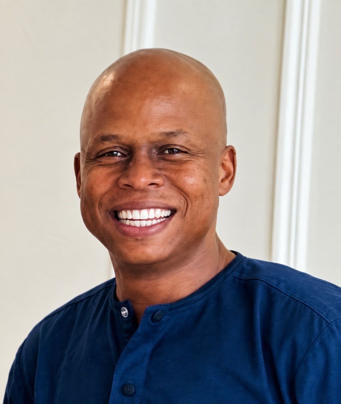 Portrait of Thapelo Kila, founder of GROUND SEED DESIGNS, smiling, in a navy shirt against a white panelled wall.