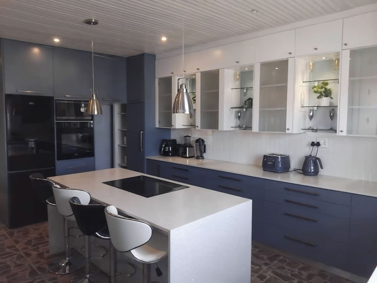 Custom navy and white kitchen by GROUND SEED DESIGNS: navy base cabinetry, white glass-fronted wall units with lit glass shelves, a white waterfall-edge island with a built-in hob and bar stools, and tall units housing the oven and microwave.