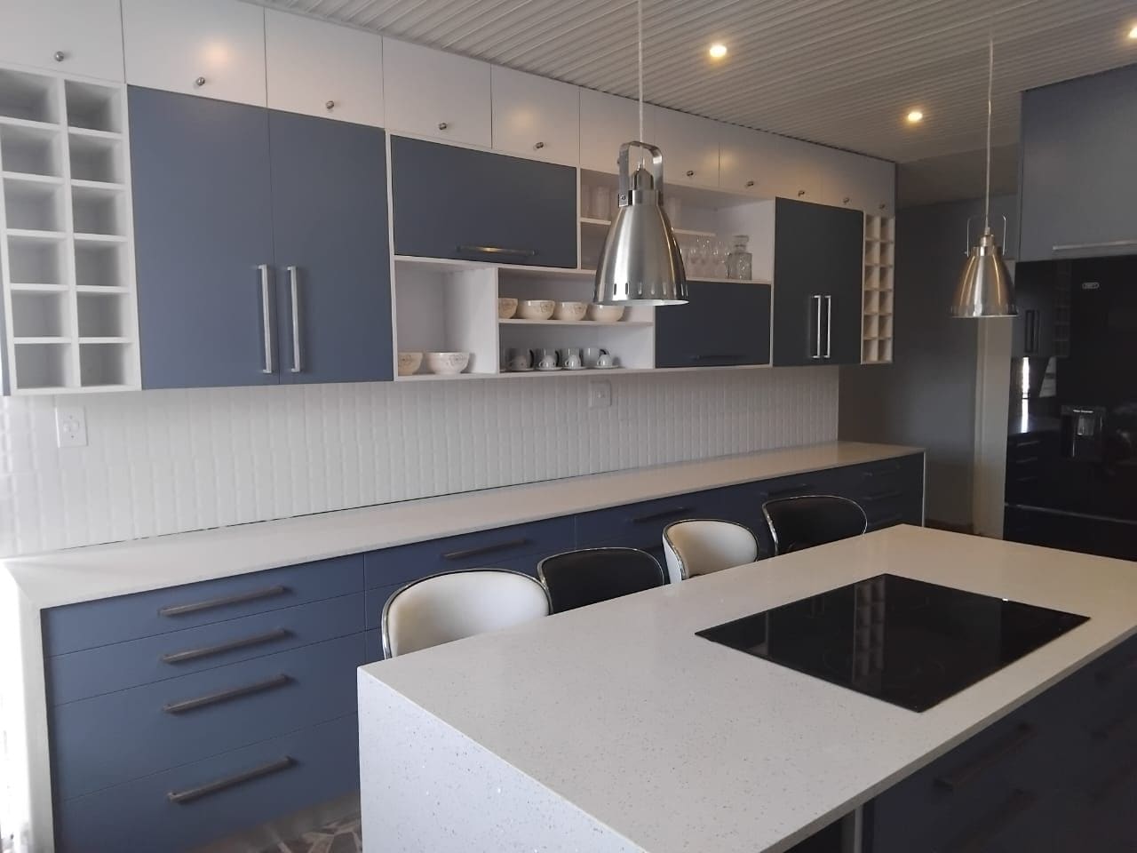 Navy and white wall units with open shelving and a cube wine rack above navy drawers, and a white island with an induction hob and seating.