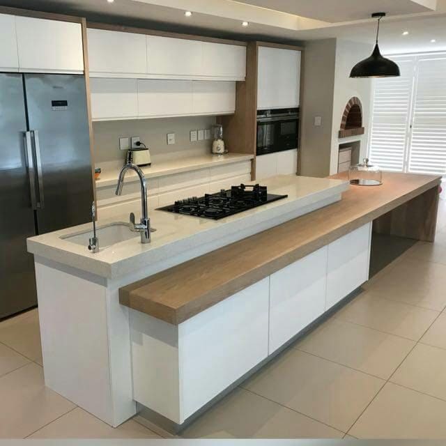 Long white island with a gas hob and sink, and a timber breakfast ledge running its full length, beside tall fridge and oven units.