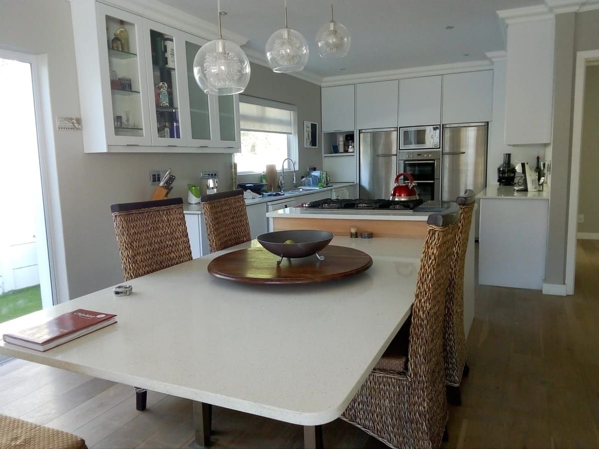 White kitchen with glass-fronted wall units and glass pendant lights over a long white table, with tall appliance units behind.