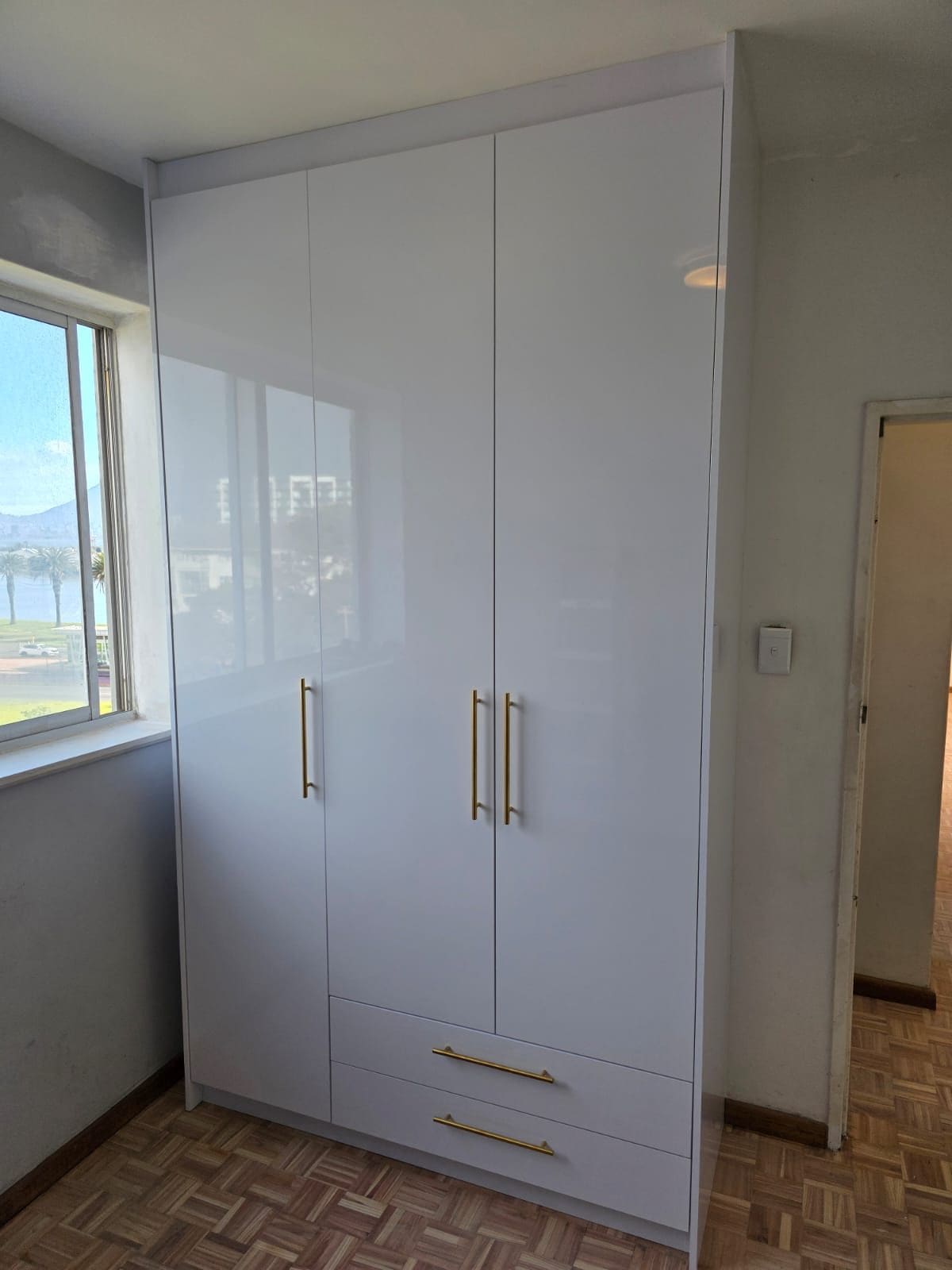 Built-in wardrobe with white high-gloss doors and two drawers beside a window, fitted floor to ceiling in a bedroom.